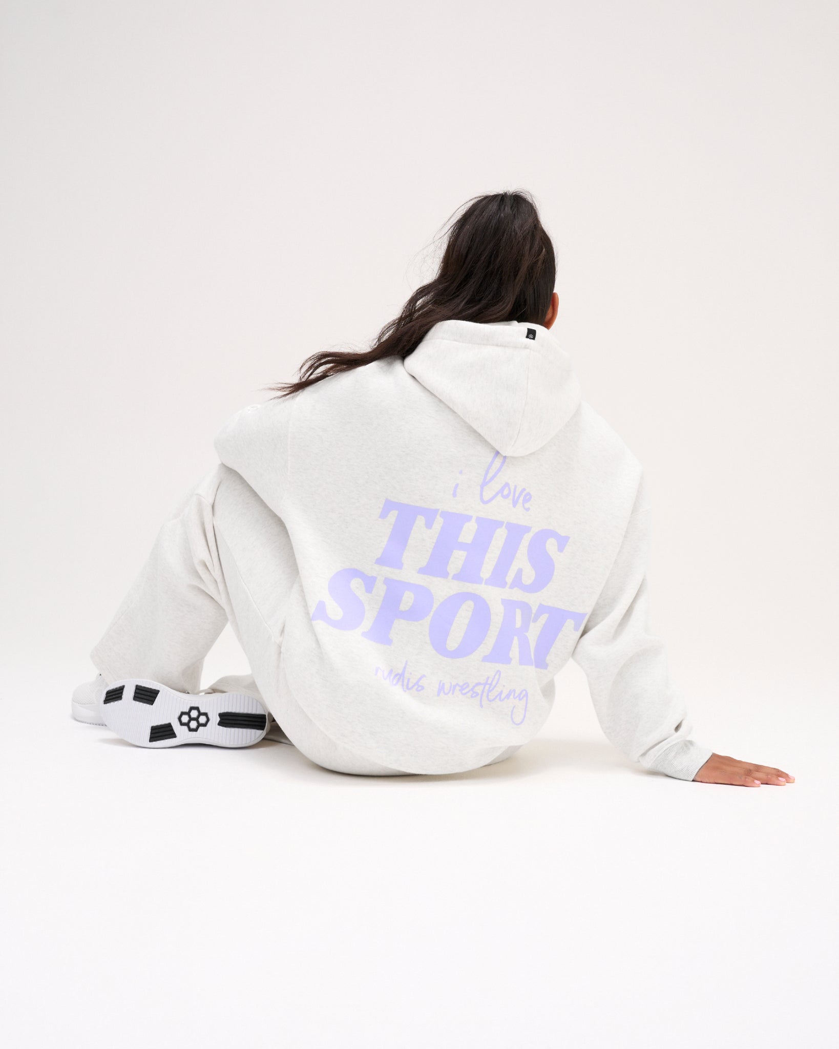 Seated female model showing the back of the RUDIS Love This Sport Heather Silver fleece hoodie with a large lavender I Love This Sport wrestling graphic.