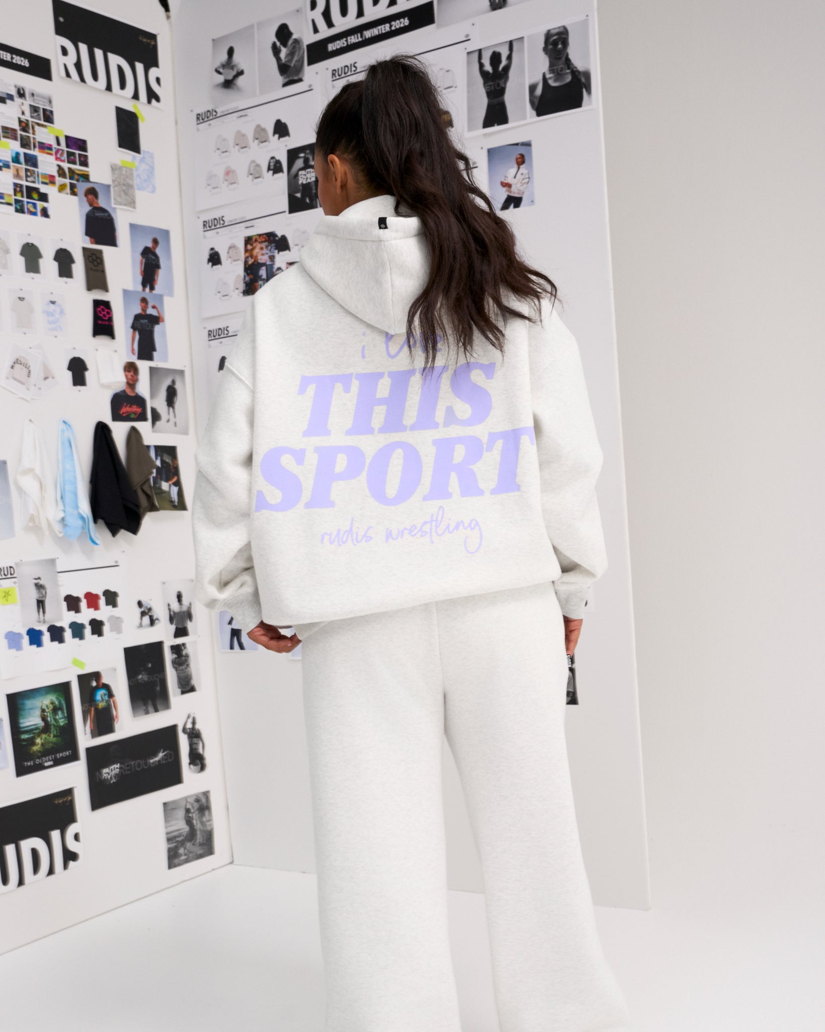 Rear view of a female model wearing the RUDIS Love This Sport Relaxed Comfort Fleece Hoodie in Heather Silver with lavender back graphics and matching sweatpants.