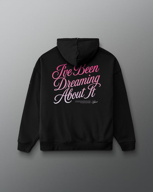 A black pullover hoodie shown from the back, featuring the phrase 'I've Been Dreaming About It' printed in a stylized pink and white script across the upper back.
