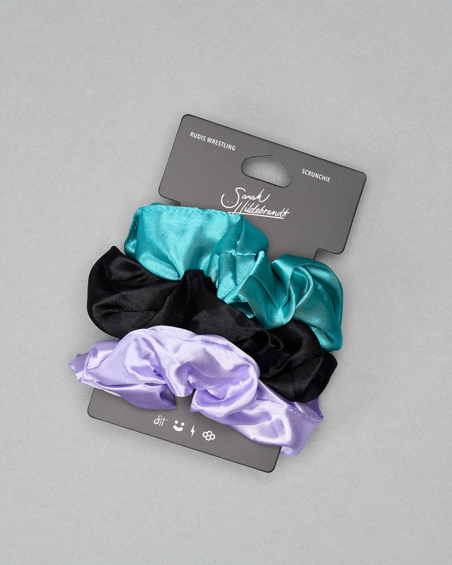Set of three satin scrunchies (black, teal, purple) on a gray background with a brand card.