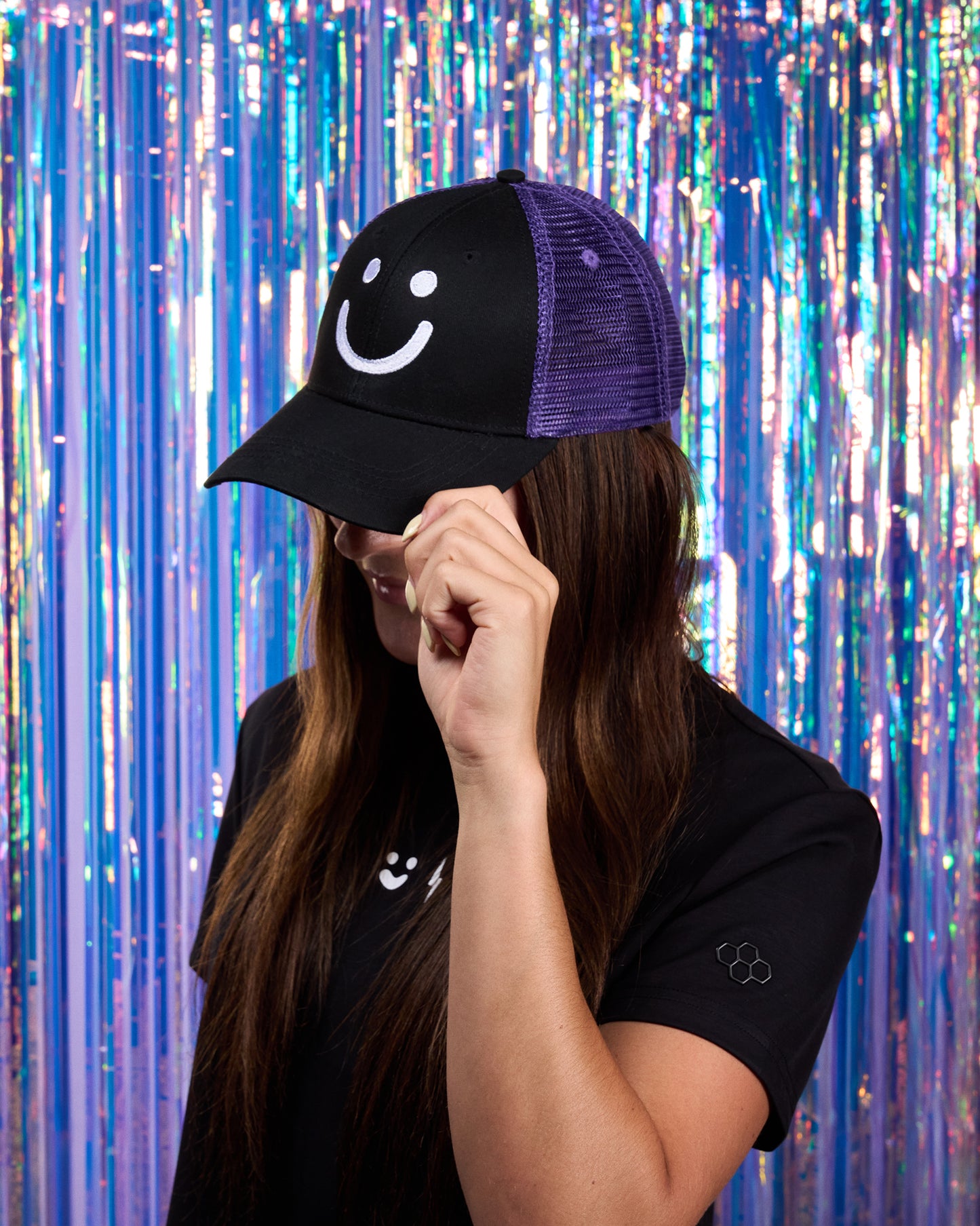 Person wearing a black cap with a smiley face design against a colorful tinsel background