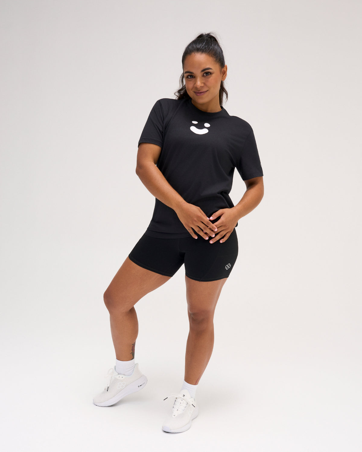 Woman modeling the black RUDIS SH Smile Elite Super Soft T-Shirt with white smile graphic, black athletic shorts, and white shoes.