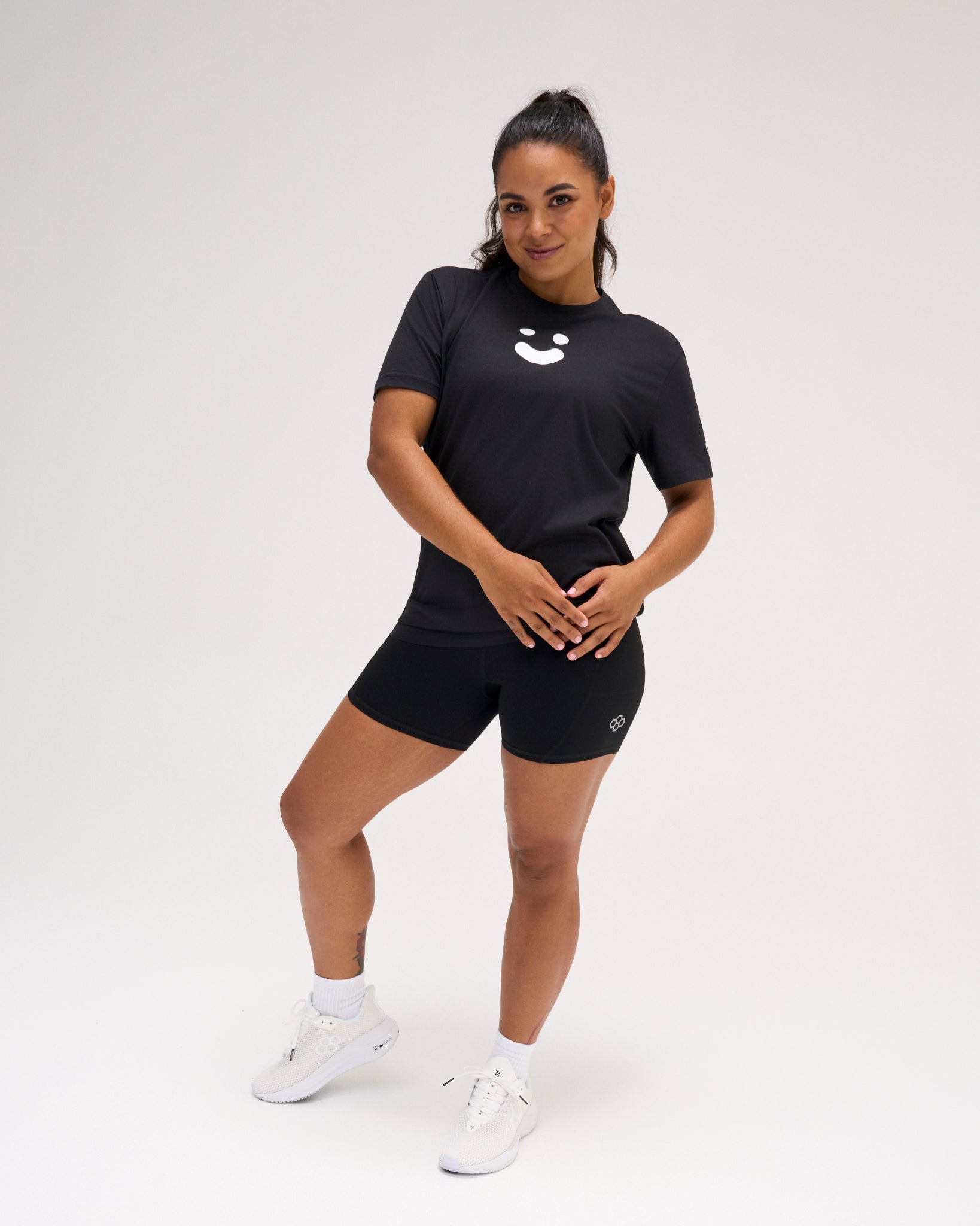 Woman modeling the black RUDIS SH Smile Elite Super Soft T-Shirt with white smile graphic, black athletic shorts, and white shoes.