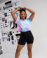 Woman modeling the RUDIS SH Dreaming Women’s Cropped Tie-Dye T-Shirt in Cotton Candy with white script graphic and black athletic shorts in a RUDIS design studio.