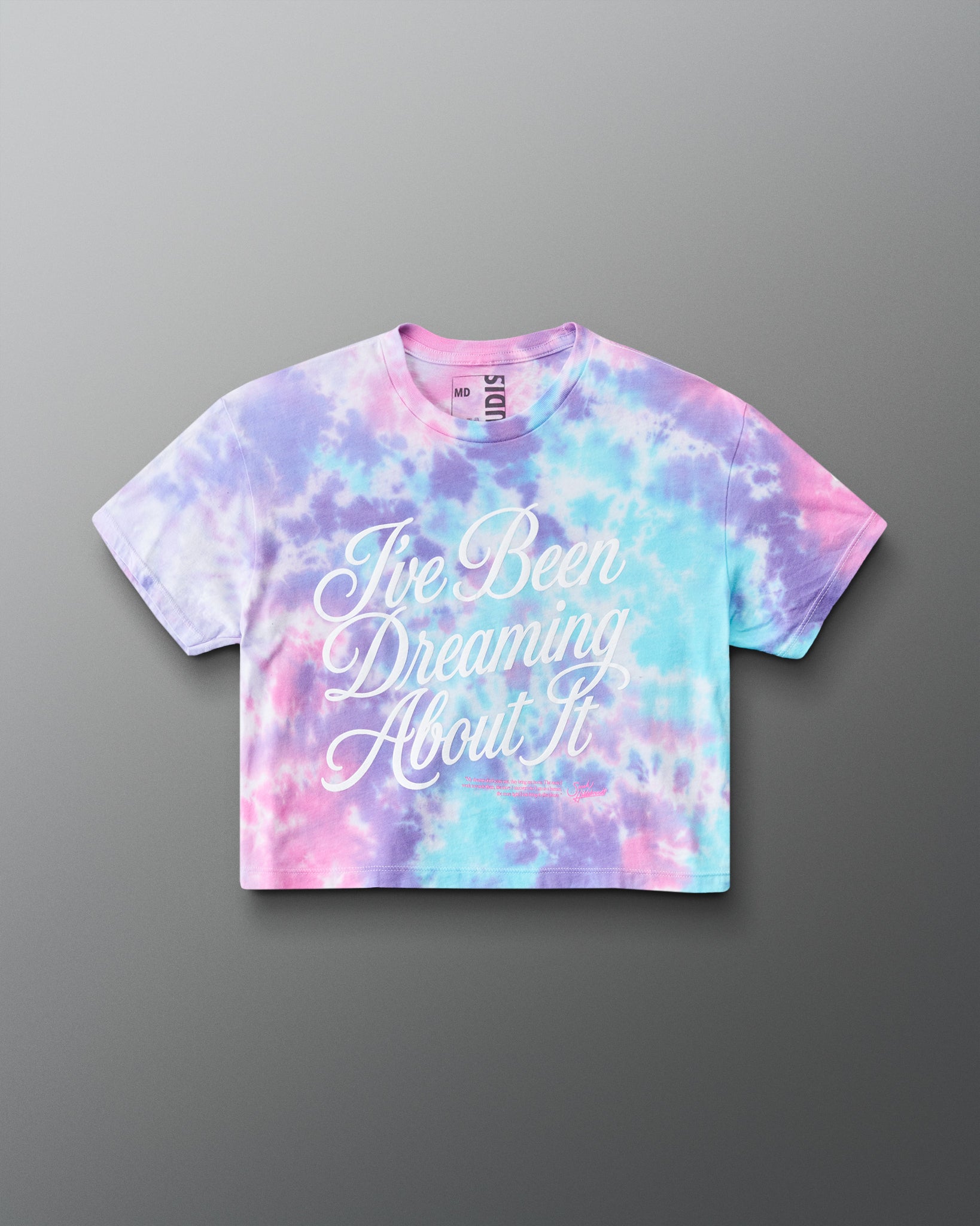 A short-sleeve crop top t-shirt featuring a pastel tie-dye pattern in pink, purple, and blue, with the phrase 'I've Been Dreaming About It' printed in white script across the front. The brand 'RUDIS' is visible on the inner neck label.