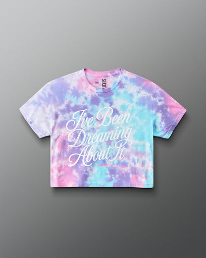 A short-sleeve crop top t-shirt featuring a pastel tie-dye pattern in pink, purple, and blue, with the phrase 'I've Been Dreaming About It' printed in white script across the front. The brand 'RUDIS' is visible on the inner neck label.