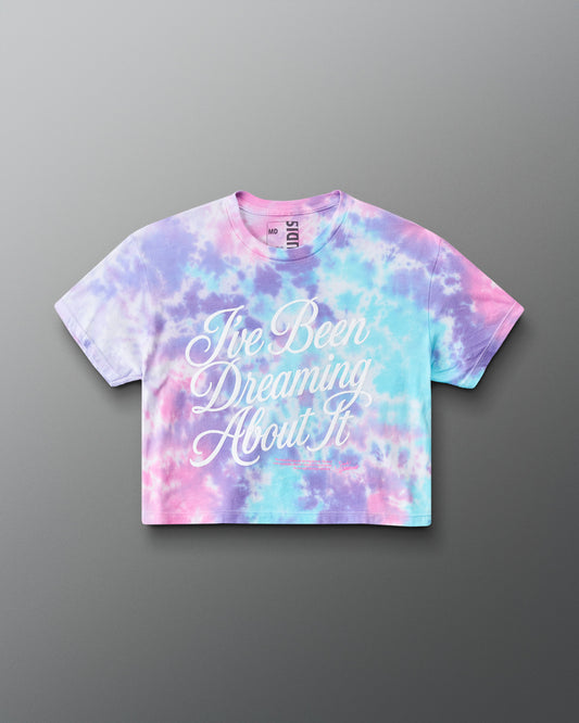 A short-sleeve crop top t-shirt featuring a pastel tie-dye pattern in pink, purple, and blue, with the phrase 'I've Been Dreaming About It' printed in white script across the front. The brand 'RUDIS' is visible on the inner neck label.