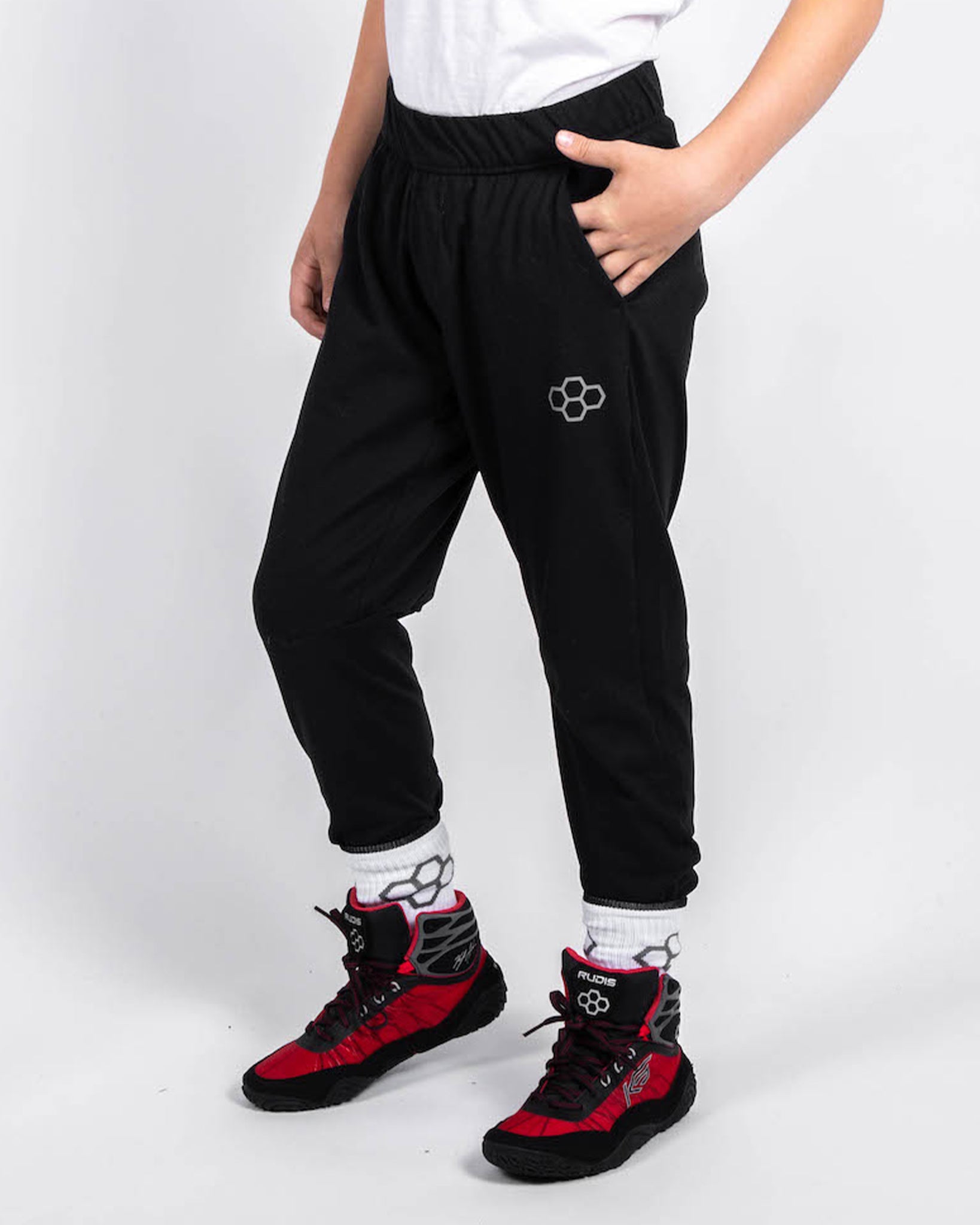 A child is standing in a casual pose wearing stylish black sweatpants with a logo paired with eye-catching red and black athletic shoes and white socks