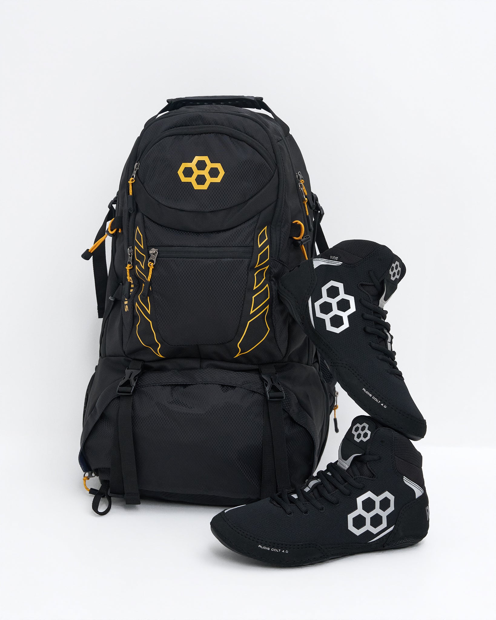 A black technical backpack with yellow logo accents, shown alongside a pair of black wrestling shoes with white and yellow logos, on a clean white background.