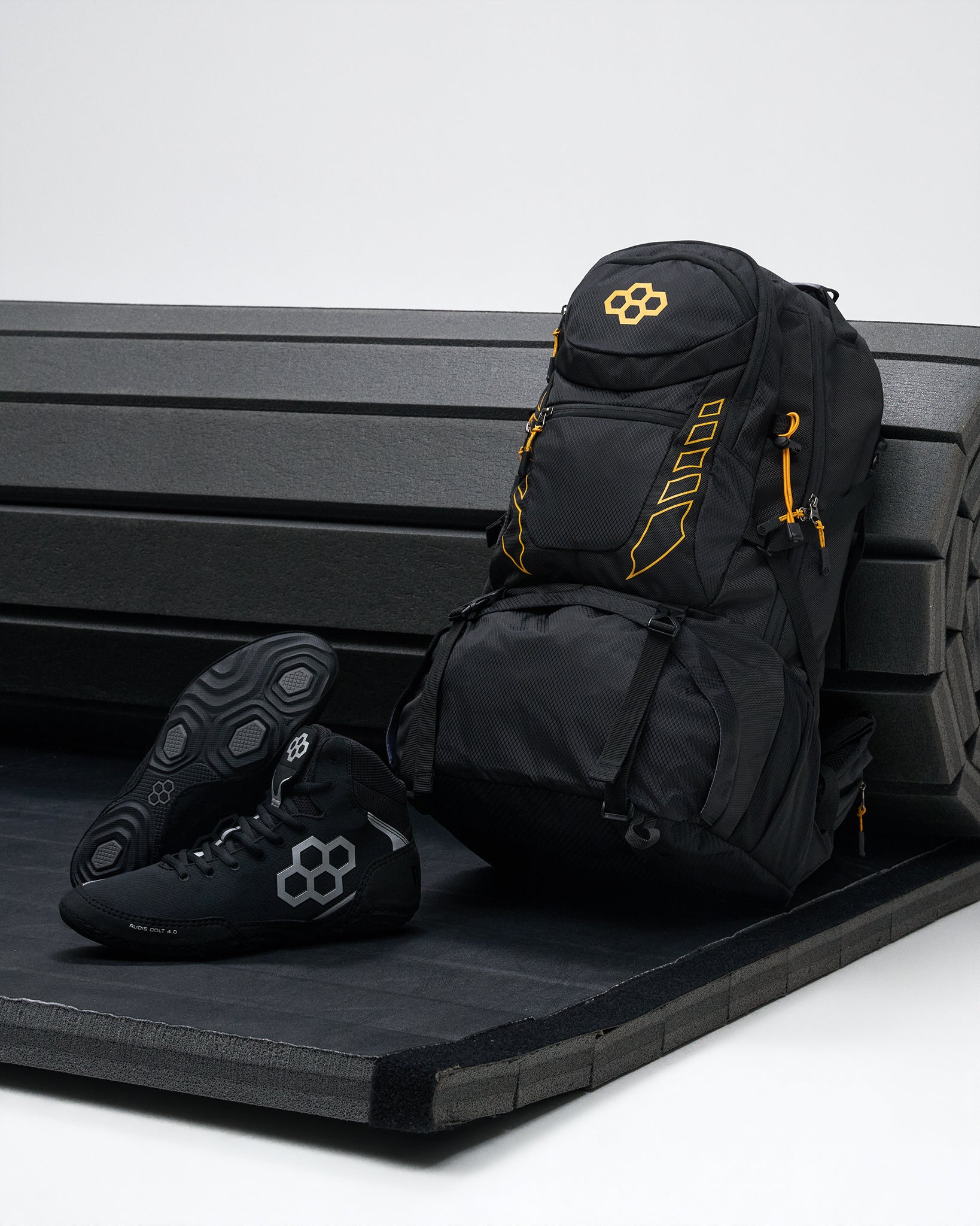 A black backpack with bright yellow accents and a matching black wrestling shoe with a gray geometric logo, displayed on a black bench against a light gray background.