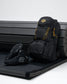 A black backpack with bright yellow accents and a matching black wrestling shoe with a gray geometric logo, displayed on a black bench against a light gray background.