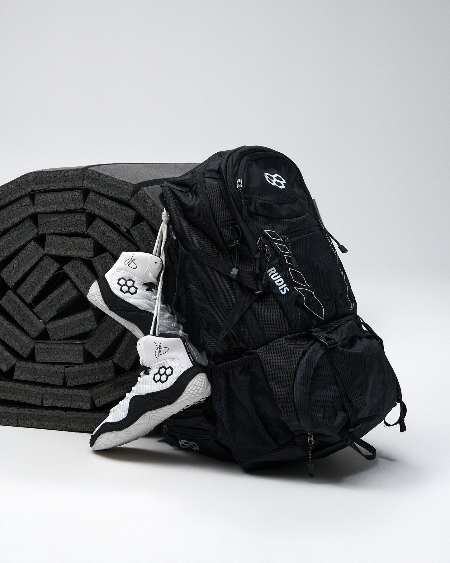 A black backpack with white logo details and a white-and-black wrestling shoes draped over its side, set against a light gray studio background with a black foam wrestling mat.