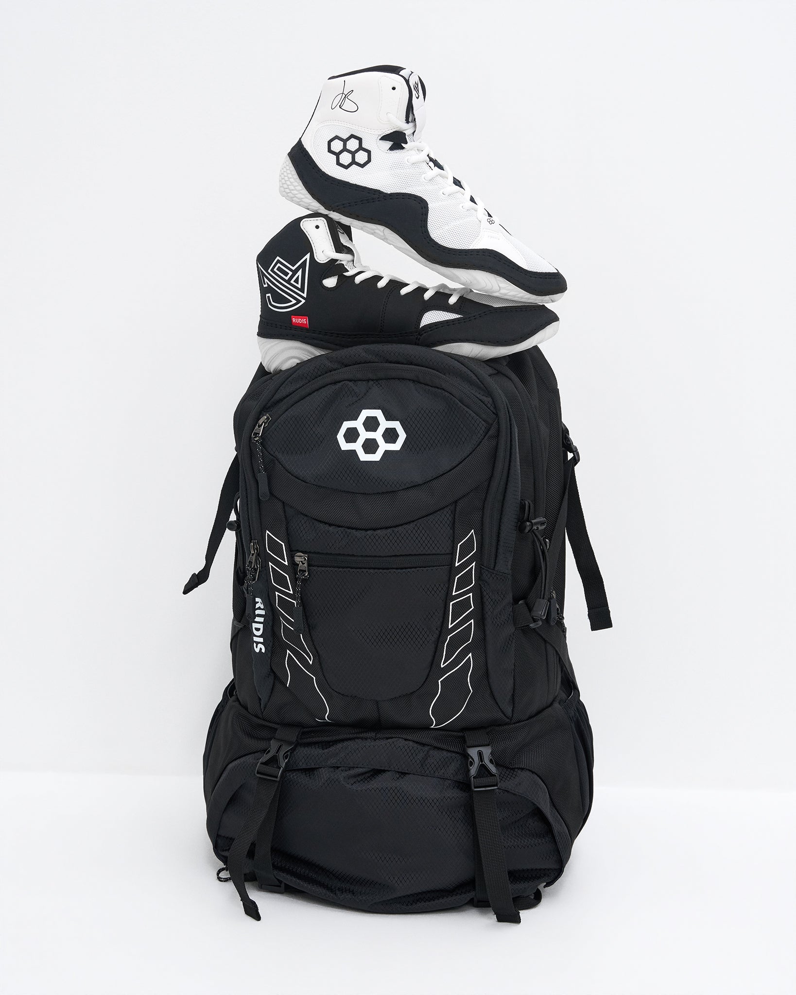 A black backpack with white graphic details and a white geometric logo on the front pocket, styled with a pair of black and white high-top wrestling shoes stacked on top, set against a plain white studio background.