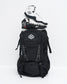 A black backpack with white graphic details and a white geometric logo on the front pocket, styled with a pair of black and white high-top wrestling shoes stacked on top, set against a plain white studio background.