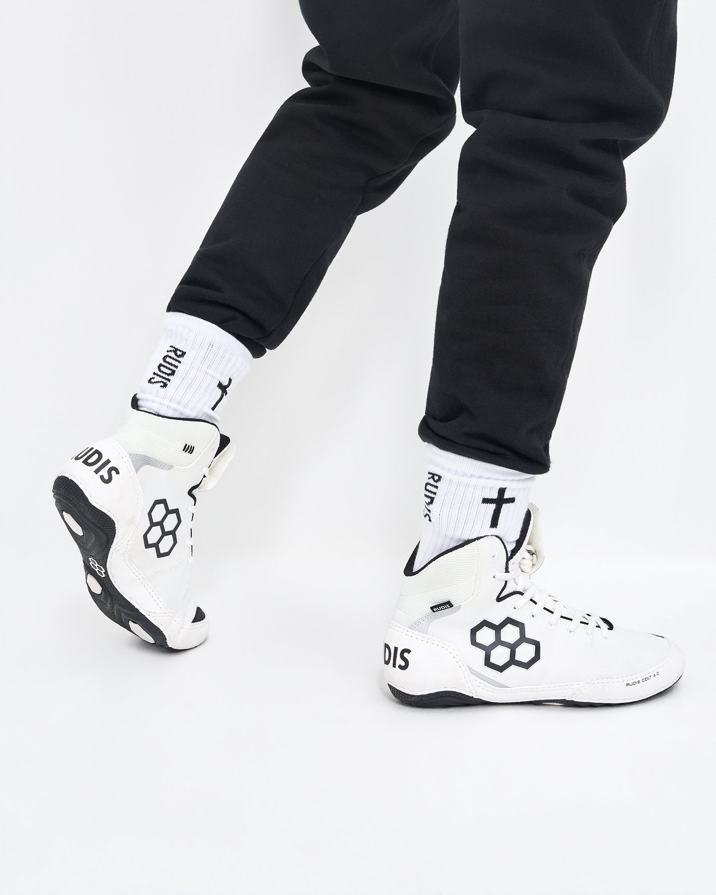 White and black high-top wrestling shoes with white crew socks featuring black text and a cross, worn with black pants against a white studio background.