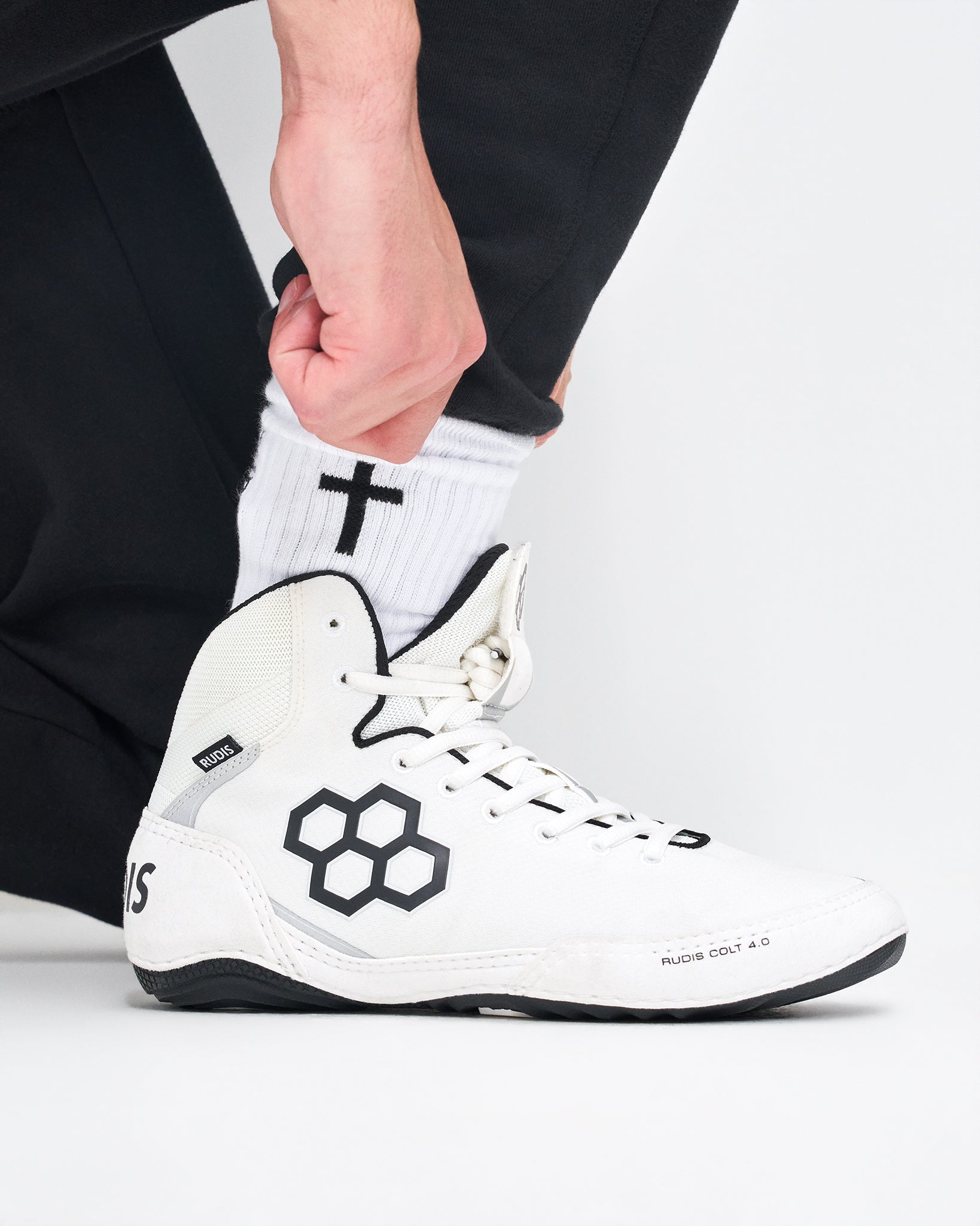 A close-up of a person's foot wearing a white high-top wrestling shoe with black accents, paired with a white crew sock featuring a black cross, set against a plain light gray background.