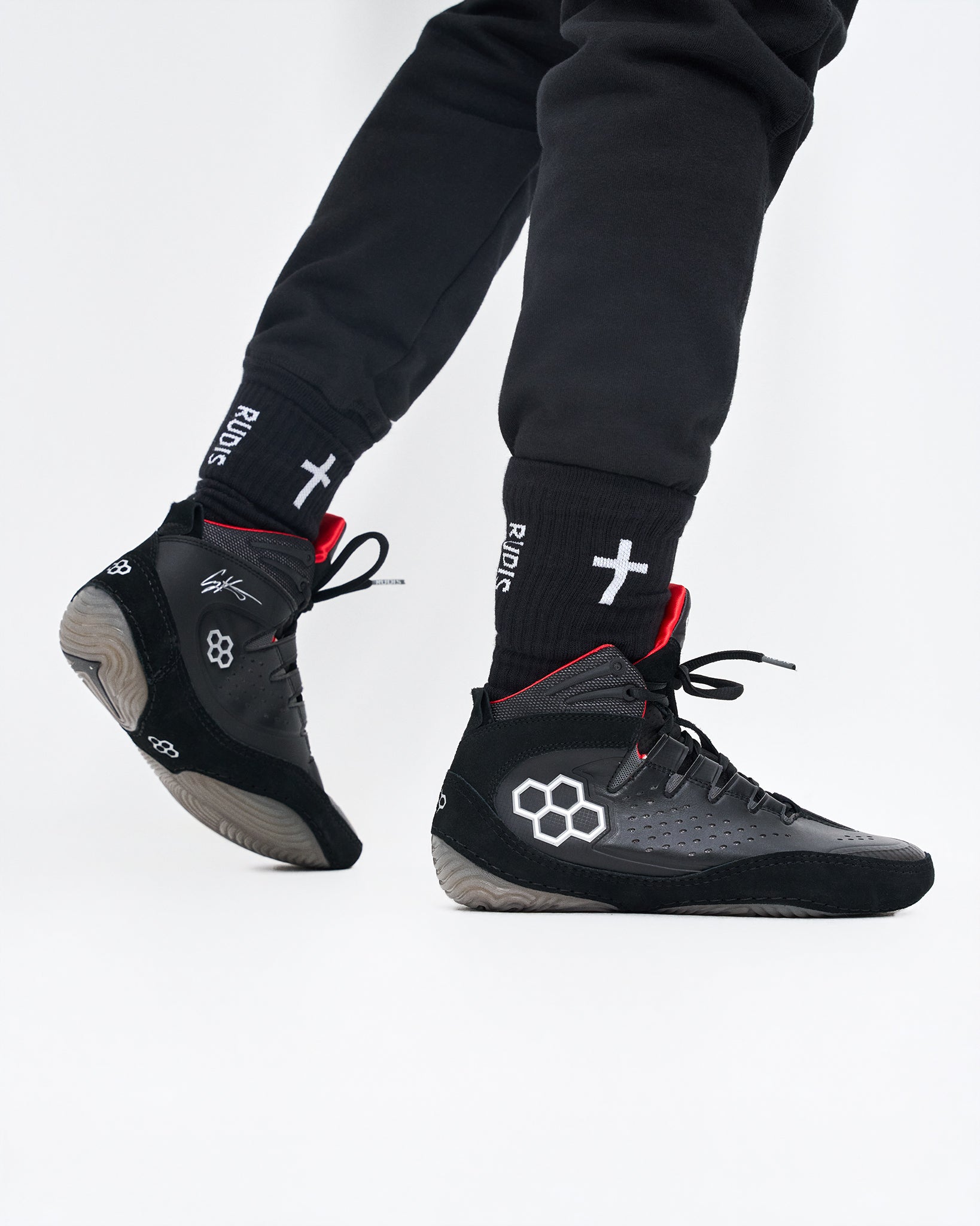 Black high-top wrestling shoe with white geometric and cross logos, worn with black socks featuring a white cross and the brand name 'RUDIS'.