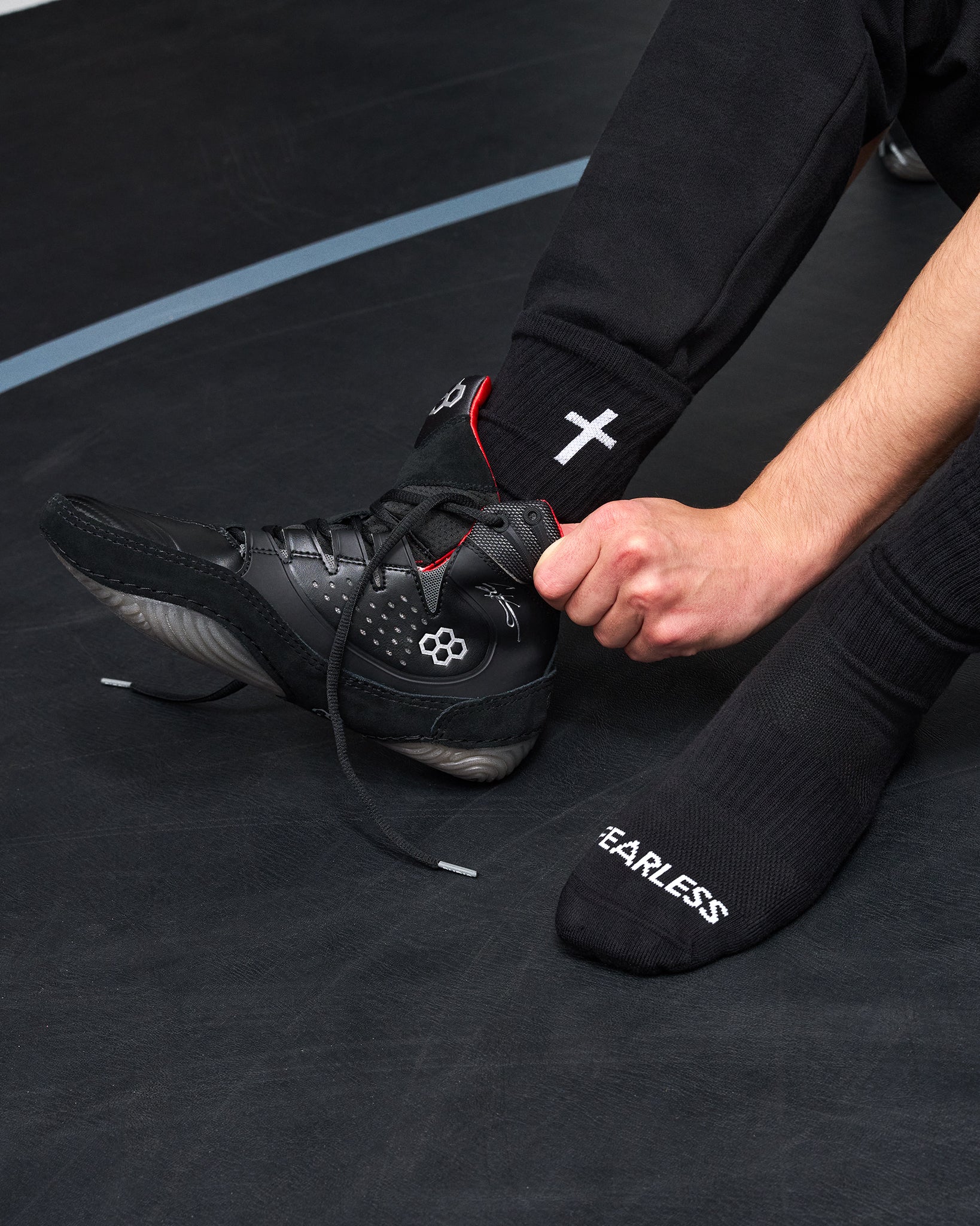 A close-up of a person's foot and hand adjusting a black wrestling shoe, wearing black crew socks with white text and logos, on a dark gym floor.