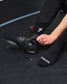 A close-up of a person's foot and hand adjusting a black wrestling shoe, wearing black crew socks with white text and logos, on a dark gym floor.