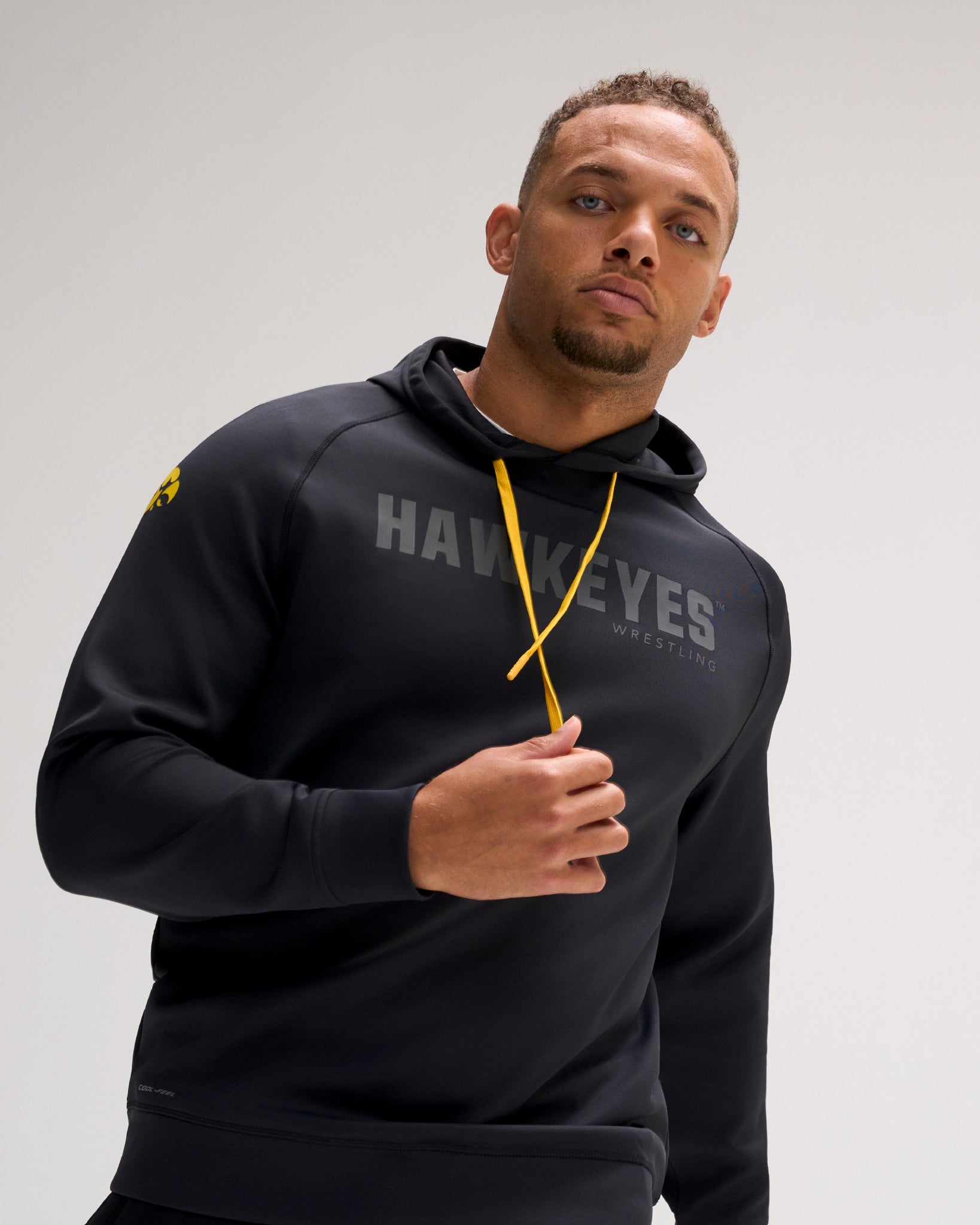 Model wearing the RUDIS Iowa Hawkeyes Cool Feel Hoodie in black with tonal Hawkeyes Wrestling chest graphic, gold drawcords, and Iowa logo on the sleeve.
