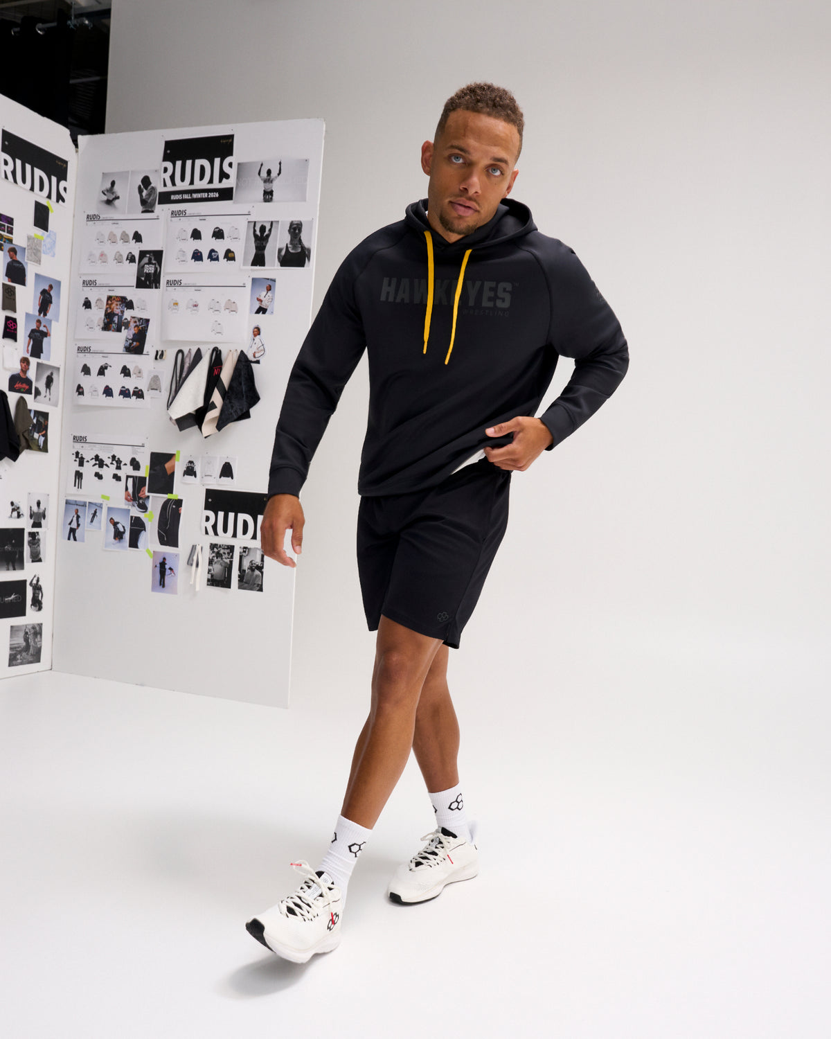 Model wearing the RUDIS Iowa Hawkeyes Cool Feel Hoodie in black with gold drawcords, paired with black athletic shorts and white training shoes.