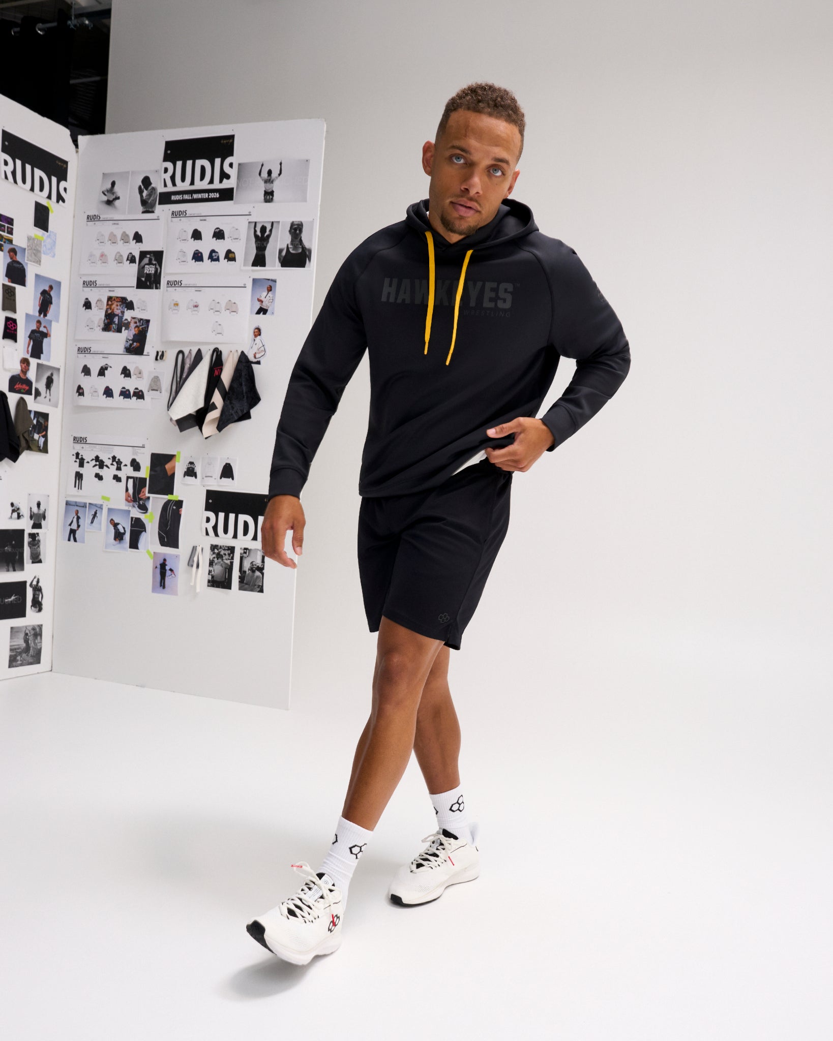 Model wearing the RUDIS Iowa Hawkeyes Cool Feel Hoodie in black with gold drawcords, paired with black athletic shorts and white training shoes.