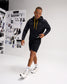 Model wearing the RUDIS Iowa Hawkeyes Cool Feel Hoodie in black with gold drawcords, paired with black athletic shorts and white training shoes.