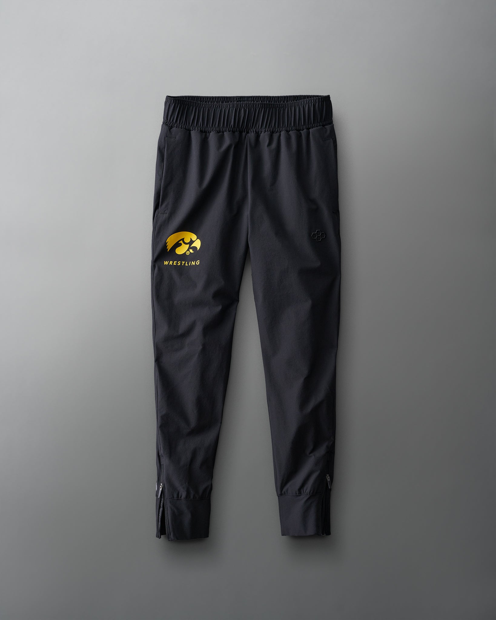 Youth athletic pants in black with a gold iowa hawkeyes wrestling logo on the left leg, laid flat on a grey background.