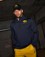 Man wearing a navy blue zip-up with a logo and yellow pants, standing against a dark background.