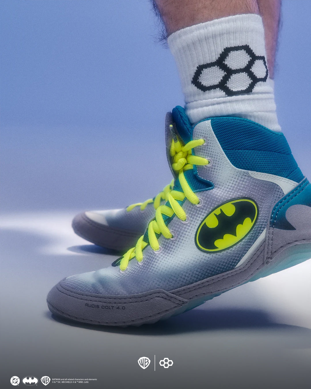 A close-up of a person's foot wearing a gray and teal high-top wrestling shoe with neon yellow laces and a large Batman logo on the side, paired with a white crew sock featuring a black geometric pattern.