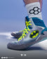 A close-up of a person's foot wearing a gray and teal high-top wrestling shoe with neon yellow laces and a large Batman logo on the side, paired with a white crew sock featuring a black geometric pattern.