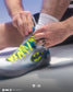 A close-up of a person tying the laces of a light gray and blue Batman-themed athletic shoe. The shoe features a prominent yellow-green Batman logo on the side, neon yellow laces, and a white sock on the ankle.