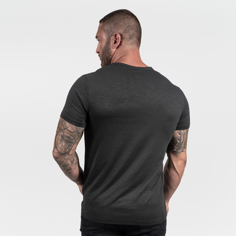 A muscular man with tattoos is seen from the back wearing a dark gray t-shirt and black jeans against a plain background