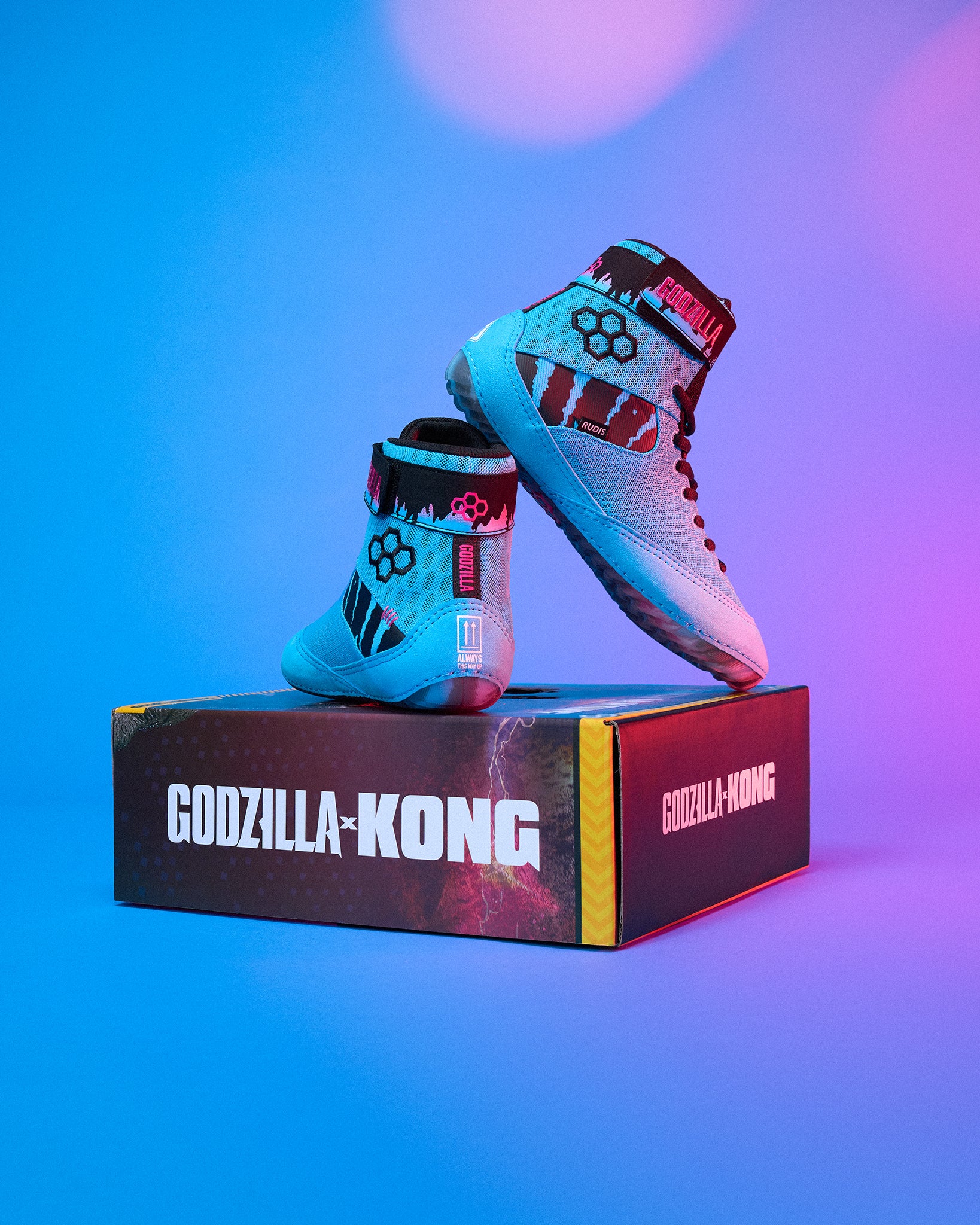 A pair of Godzilla-Kong themed high-top wrestling shoes in light blue, black, and red, displayed on top of their branded black and red shoebox against a vibrant blue and purple gradient background.