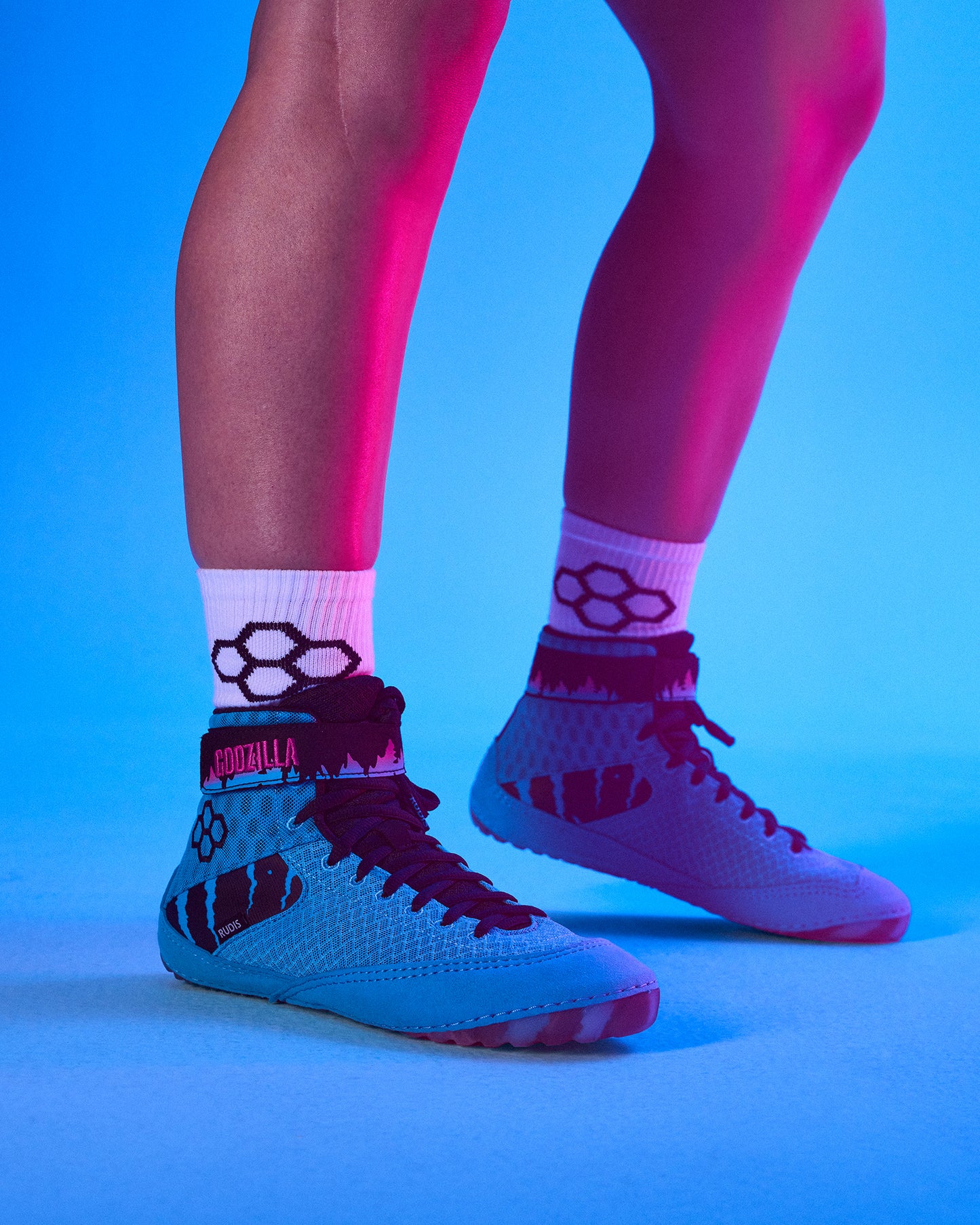 A close-up of a person's legs wearing light blue high-top wreslting shoes with dark red laces and socks, set against a vibrant blue and pink studio background.