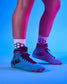 A close-up of a person's legs wearing light blue high-top wreslting shoes with dark red laces and socks, set against a vibrant blue and pink studio background.
