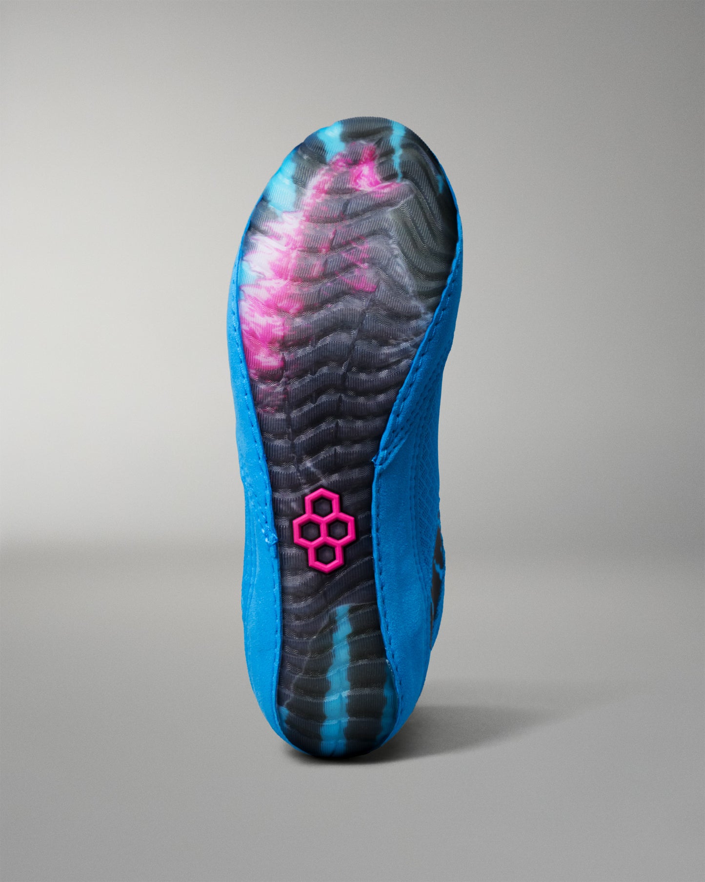 A single wrestling shoe shown from the sole view, featuring a vibrant blue upper, a black and blue patterned outsole, and a bright pink hexagonal logo near the heel, set against a neutral gray background.