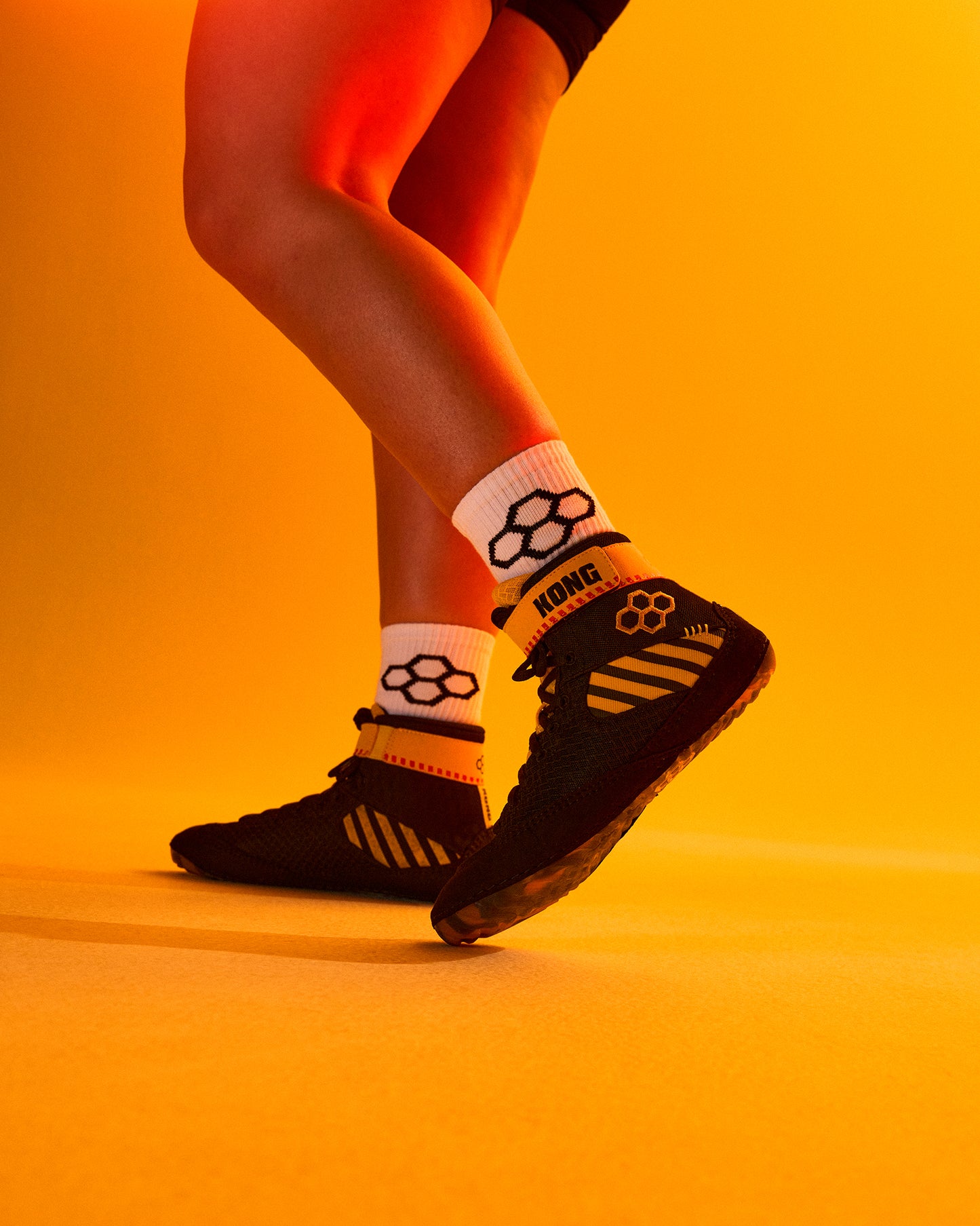 A close-up lifestyle shot of a person's legs wearing black RUDIS wrestling shoes and white crew socks with black logos, set against a warm orange-yellow studio background.
