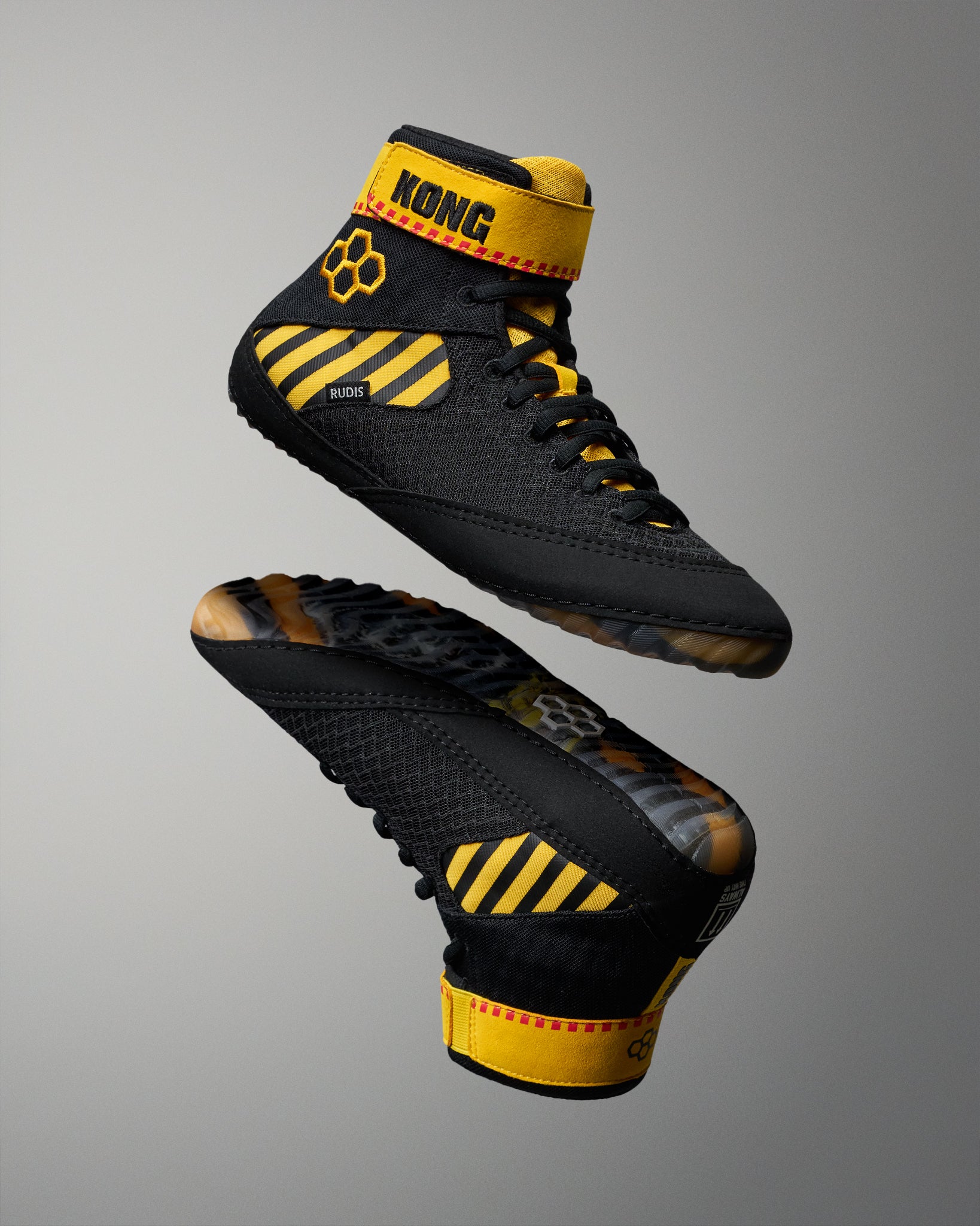 A pair of black wrestling shoes with bold yellow accents, including a yellow ankle collar, diagonal yellow stripes on the side, and a paw print logo, shown against a neutral gray gradient background.