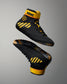 A pair of black wrestling shoes with bold yellow accents, including a yellow ankle collar, diagonal yellow stripes on the side, and a paw print logo, shown against a neutral gray gradient background.