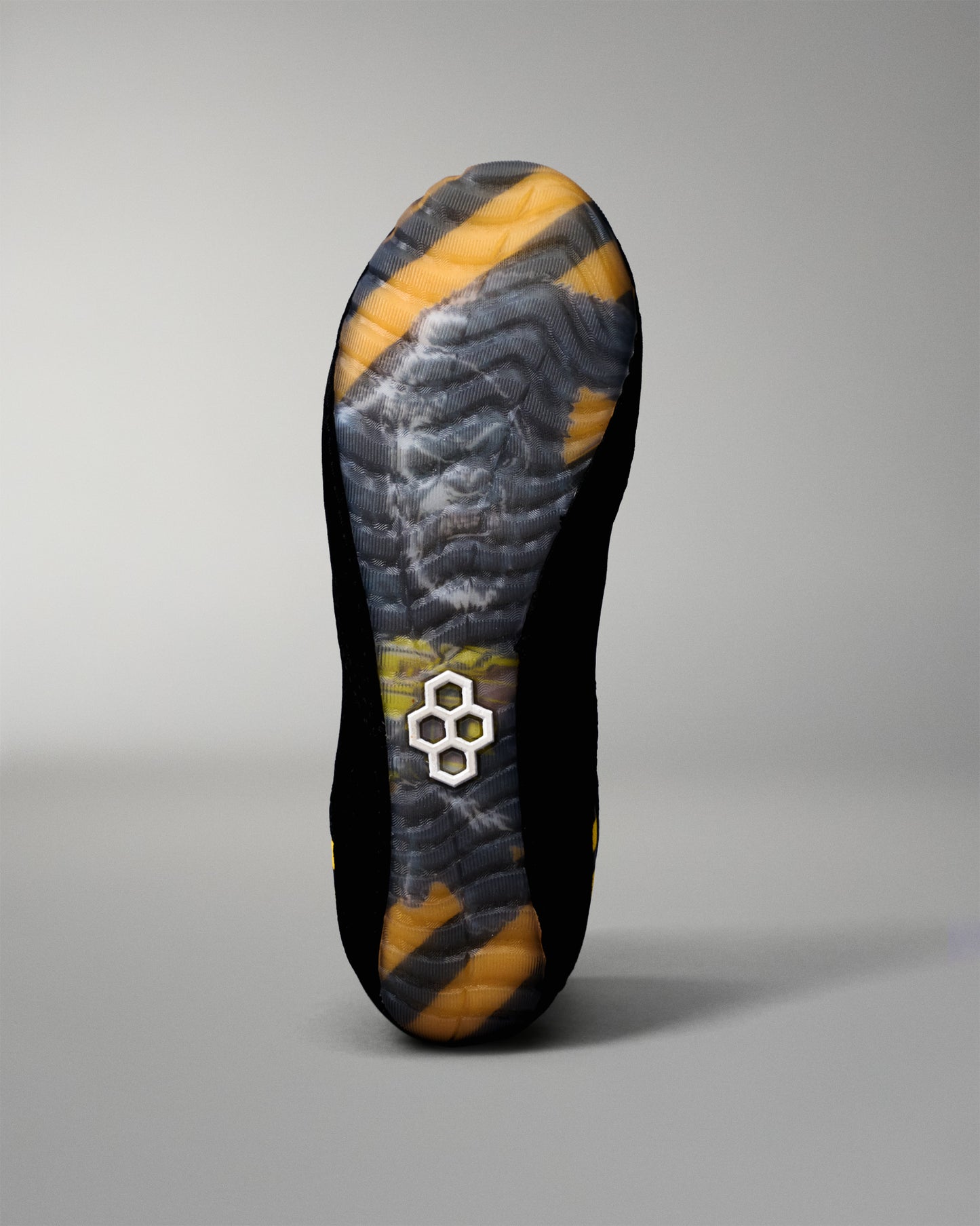 The outsole of a black wreslting shoe with a yellow and gray abstract pattern and a small white geometric logo near the center, shown against a neutral gray background.