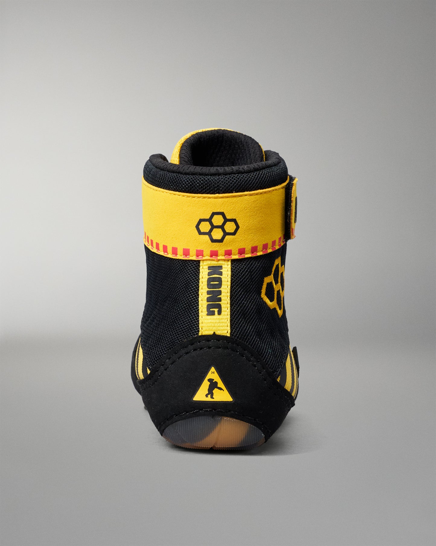 A single high-top wrestling shoe shown from the back on a neutral gray studio background. The shoe is primarily black with bold yellow accents, including a wide yellow strap around the ankle, a vertical yellow tab, and yellow graphic details.