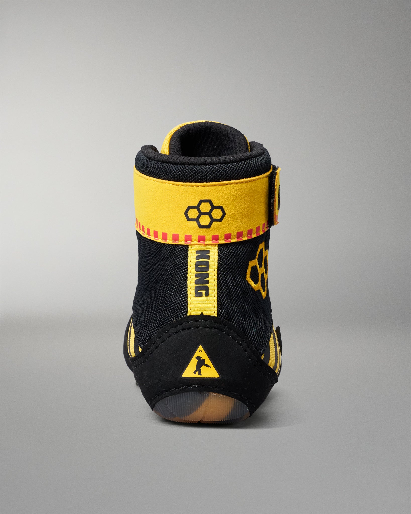 A single high-top wrestling shoe shown from the back on a neutral gray studio background. The shoe is primarily black with bold yellow accents, including a wide yellow strap around the ankle, a vertical yellow tab, and yellow graphic details.