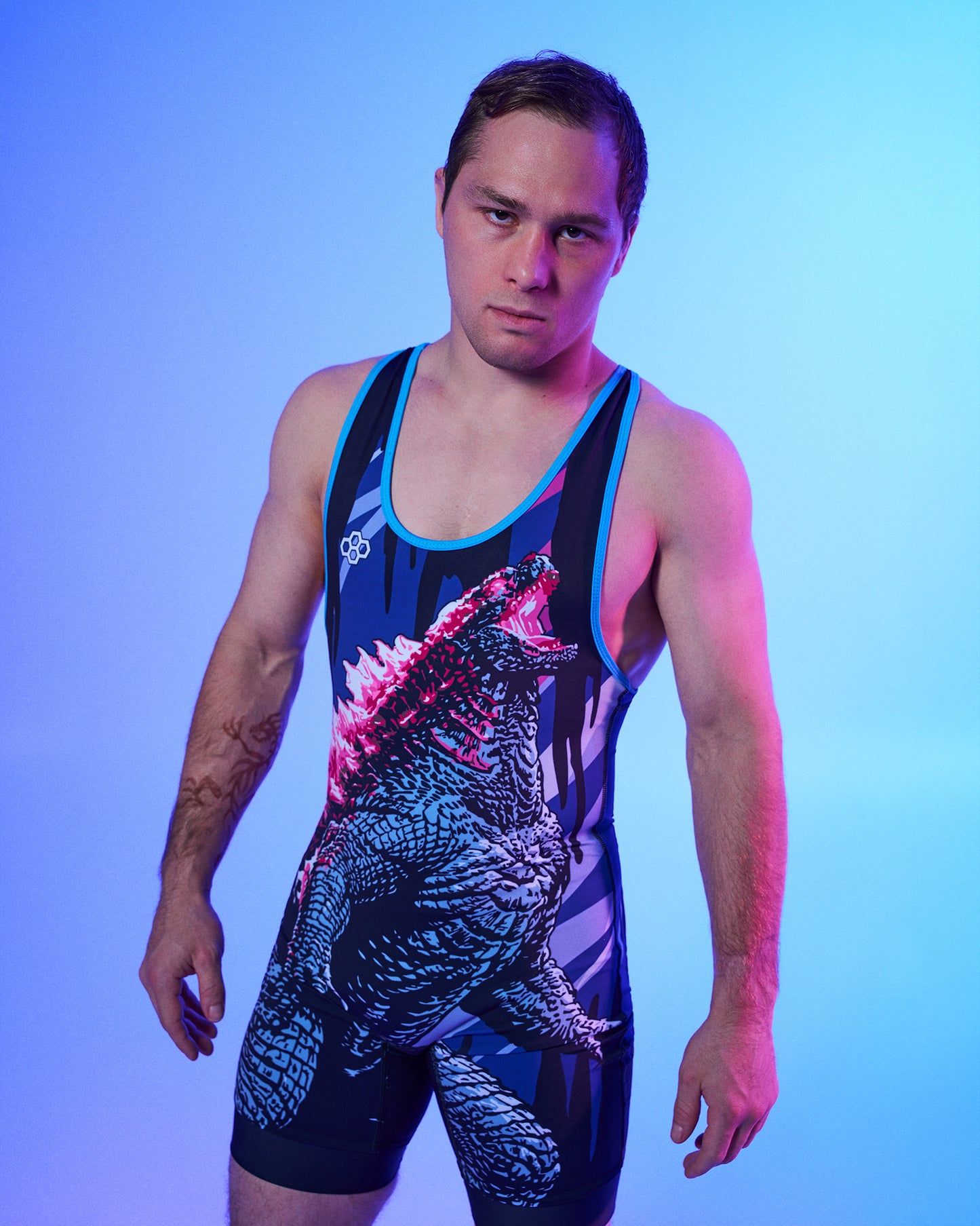 A man modeling a sleeveless wrestling singlet with a Godzilla graphic print, set against a blue and purple gradient studio background.
