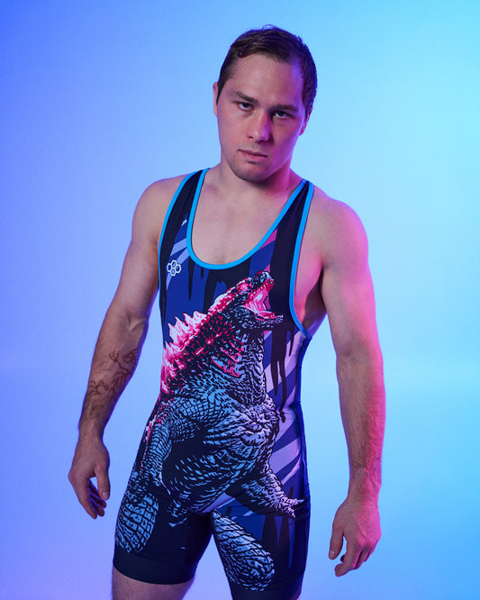 A man modeling a sleeveless wrestling singlet with a Godzilla graphic print, set against a blue and purple gradient studio background.