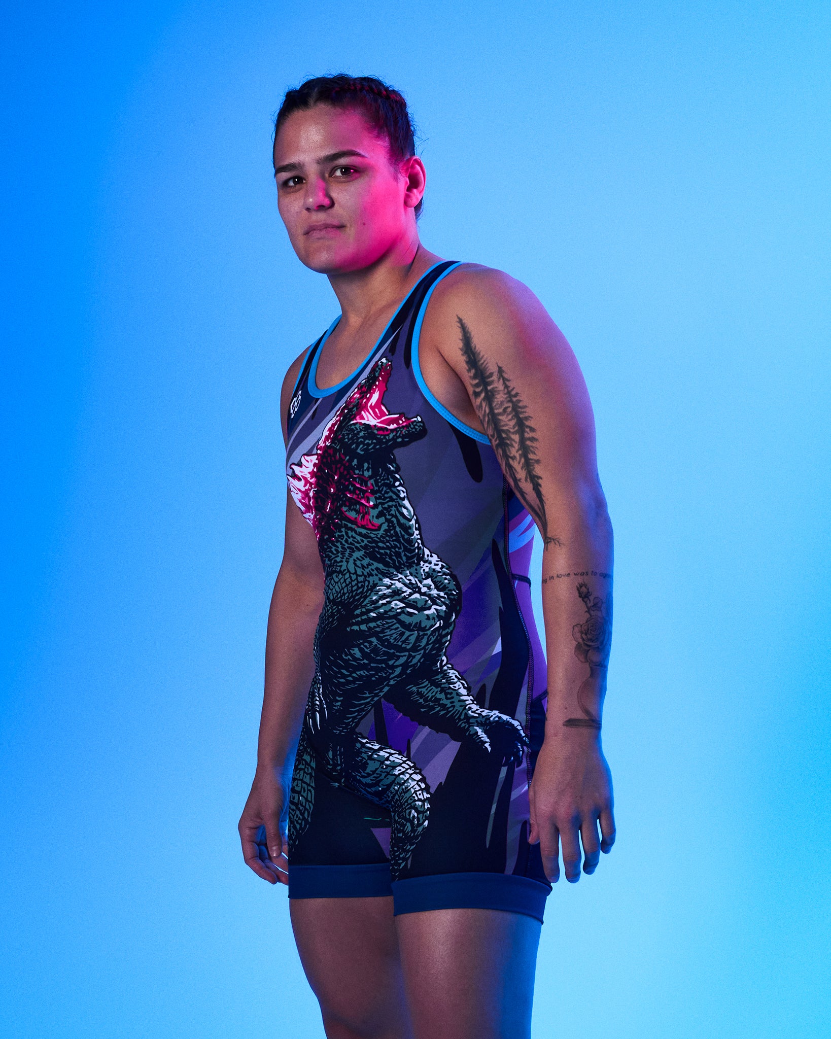 A woman modeling a sleeveless wrestling singlet with a large Godzilla graphic on the front, photographed against a bright blue studio background.