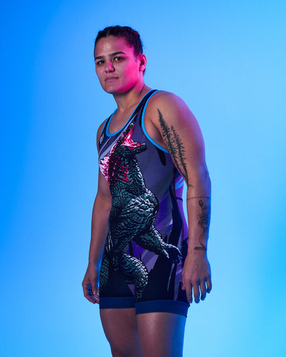 A woman modeling a sleeveless wrestling singlet with a large Godzilla graphic on the front, photographed against a bright blue studio background.