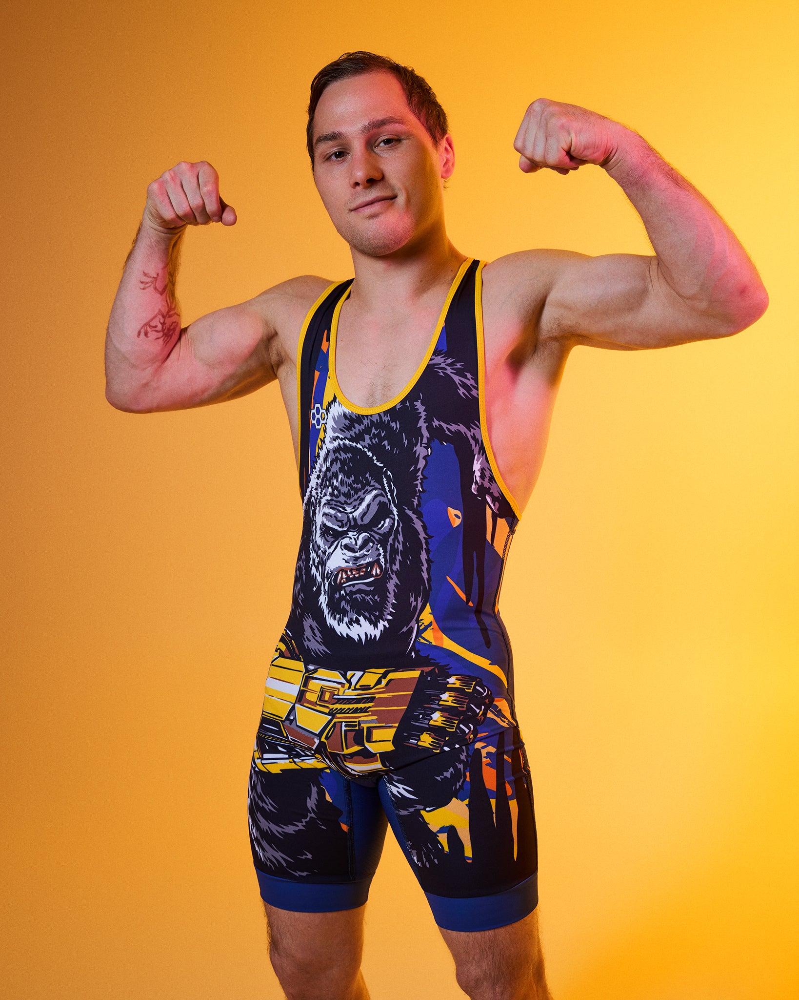 A man posing in a graphic wrestling singlet featuring a King Kong design and yellow accents, flexing his biceps against a yellow studio background.