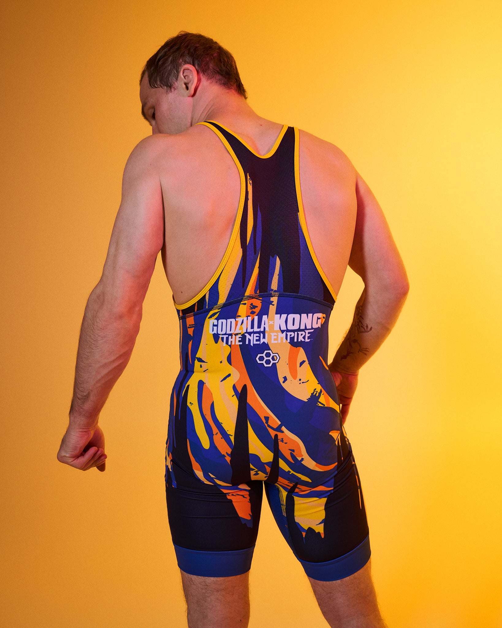 A man wearing a King Kong wrestling singlet, shown from behind against a solid yellow studio background. The singlet features a dark navy blue base with vibrant yellow and orange flame-like patterns and white text.