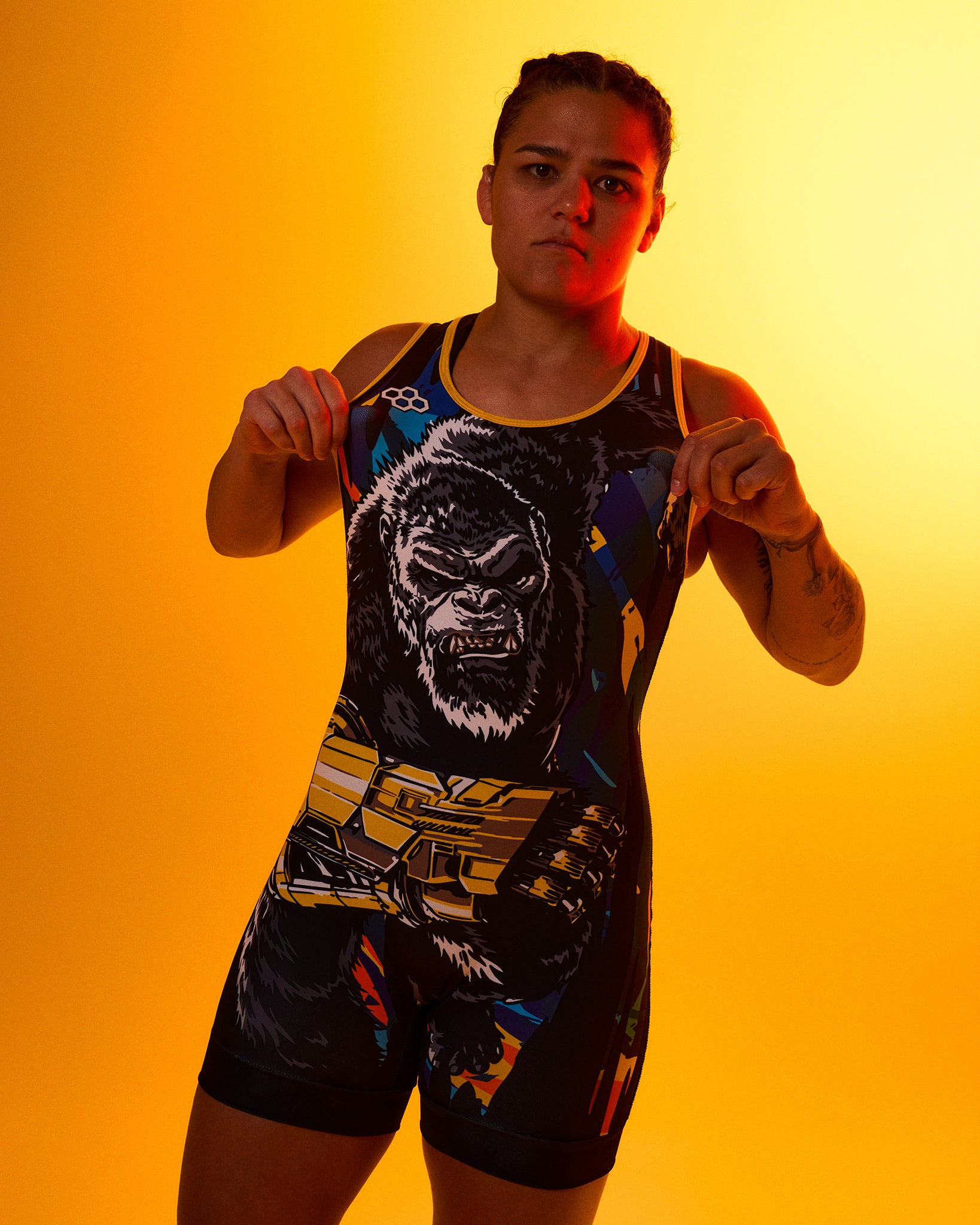 A woman modeling a black wrestling singlet with a large King Kong graphic and gold/yellow accents, posed against a warm orange-yellow studio background.